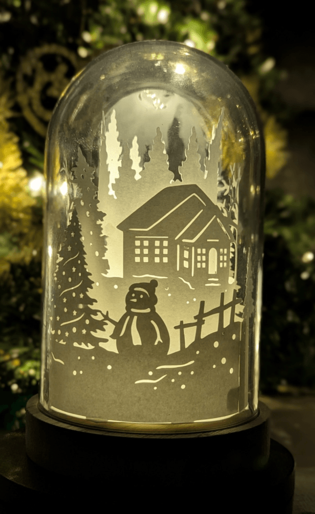 Veilleuse artisanale en papier sous cloche, scène d’hiver avec maison enneigée et bonhomme de neige, création lumineuse poétique faite main à Lille.